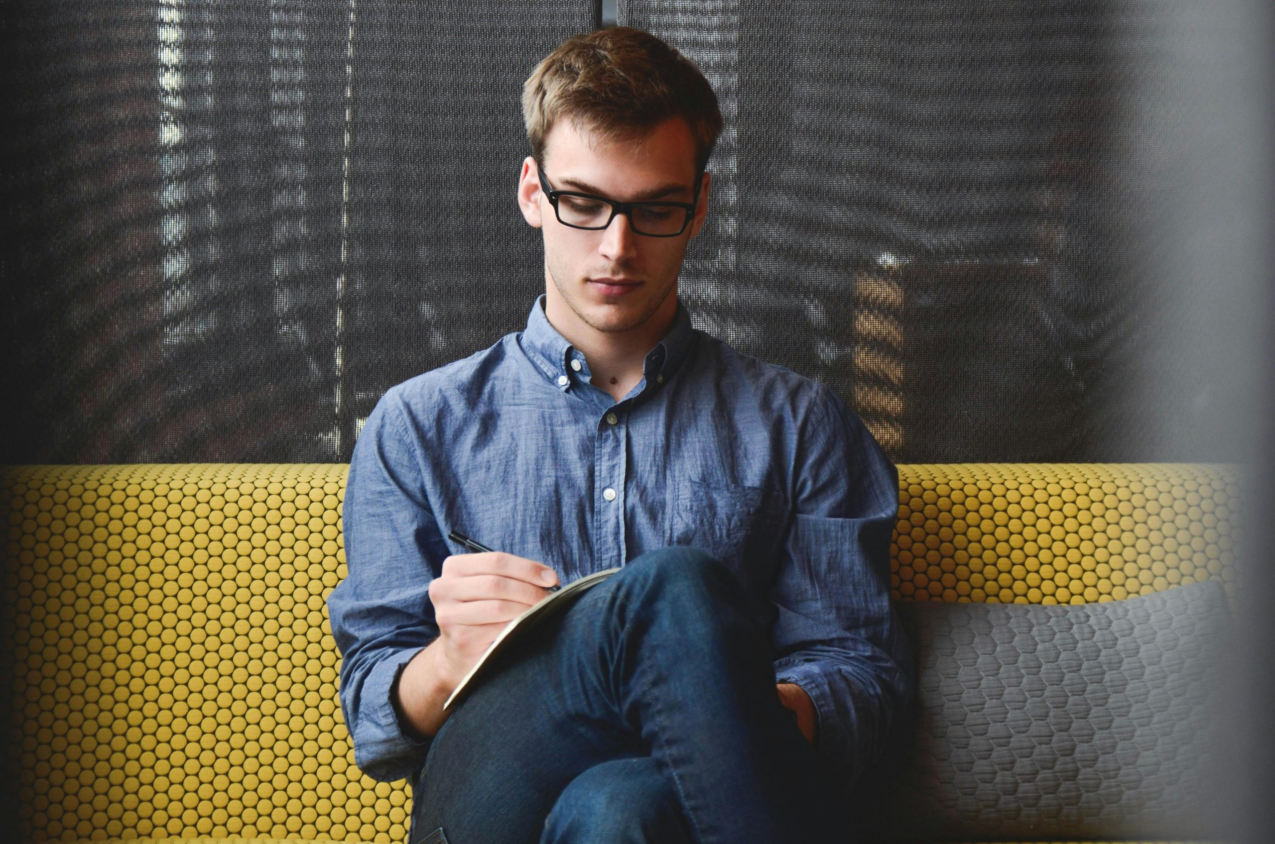 Über uns A young man in glasses writes in a notebook while sitting on a stylish couch indoors.